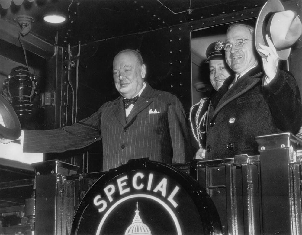 Churchill, Truman, Major General Harry Vaughan (back) wave to crowds from Presidential train car en route to Fulton